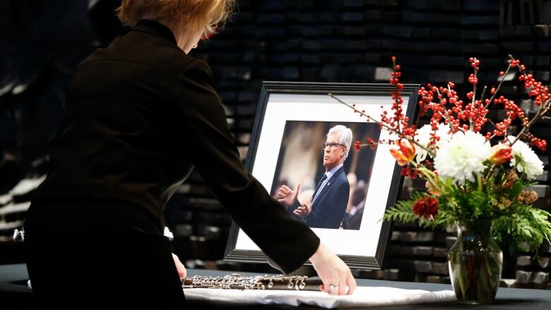 A woman dressed in black places an oboe in front of a portrait of Jim Carr on a table, with flowers in a vase to the side of the photo.