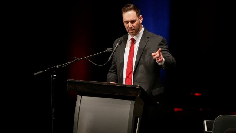 A man in a dark suit and a red tie points as he speaks at a podium.