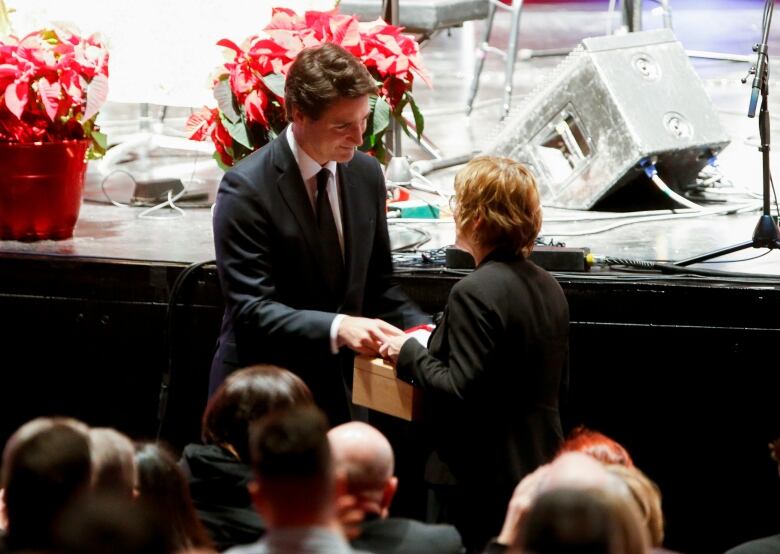 A man in a suit smiles as he hands a folded flag in a box to a woman in a dark blazer, as they stand before a crowd of people in front of a stage.