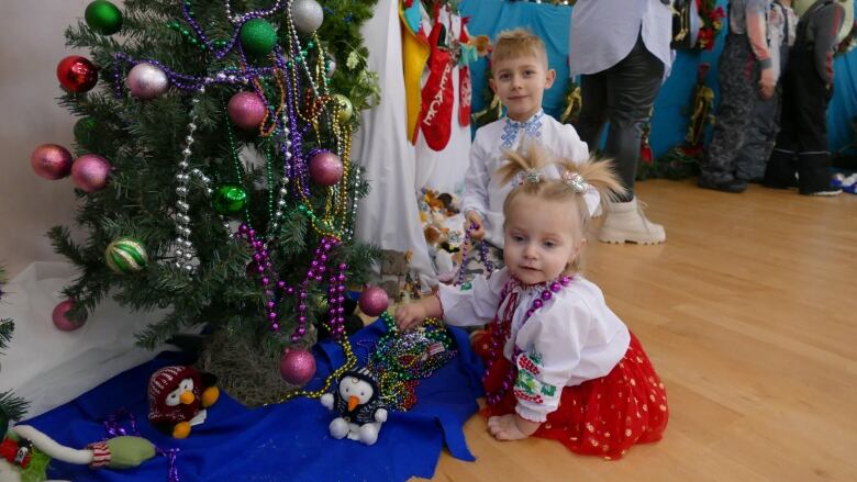 A young boy around 4-years-old, and a girl around 2-years-old sit on the floor next to a Christmas tree.