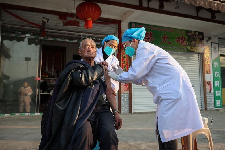 An elderly man outside with his left sleeve rolled up receives a needle from two people in white coats and blue face masks.