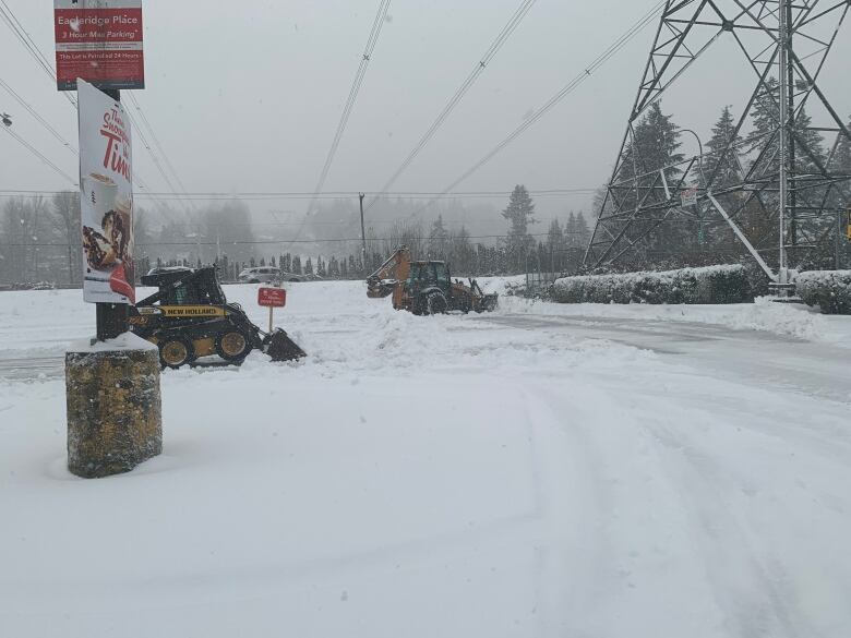 Two yellow utility vehicles shovel snow near a transmission tower.