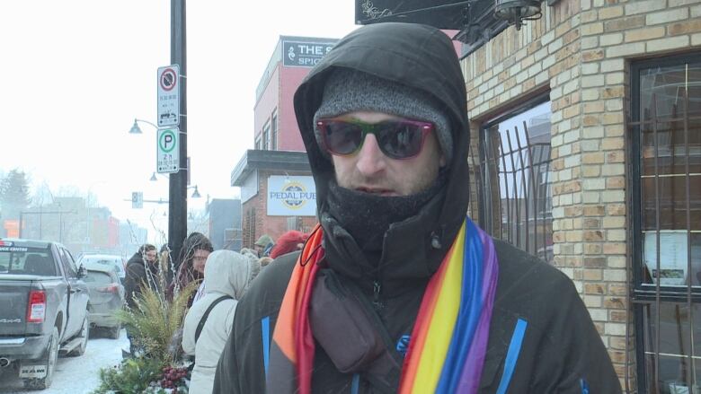 A man stands outside wearing black winter attire, sunglasses, with a rainbow flag draped around his neck. 