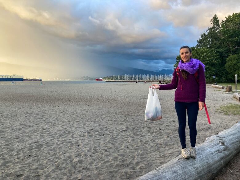 A woman stands on a log on a beach in Vancouver with the shoreline and water behind her.