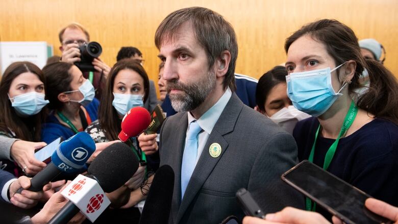 Steven Guilbeault, the minister of Environment, is surrounded by journalists, who are holding up microphones and recorders. Most of the reporters are wearing masks.