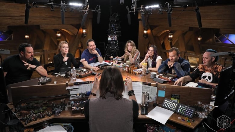 Eight people sit around a table set up with cameras and microphones playing a game of Dungeons and Dragons.