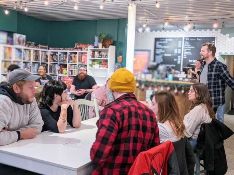 A group of people sit at tables at a board game caf, listening to the store owner. 