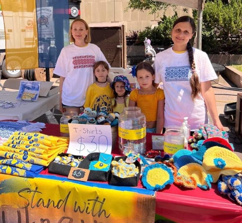 Two women and three children stand in front of colourful items such as Tshirts and hats being sold to raise money for Angels of Prince Edward Island. 