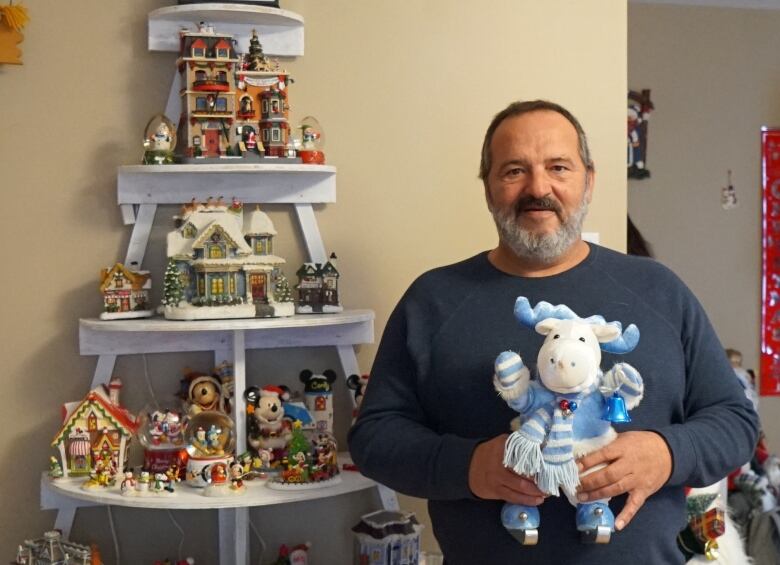 Austin Taylor stands in front of a white shelf decorated with various Christmas figurines. He is holding a blue and white moose called Mark the Moose. 