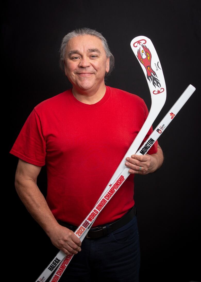 Lorne Julien a Mi'kmaw man in a red shirt hold up two hockey sticks with his designs, a speeding eagle with an orange heart. 