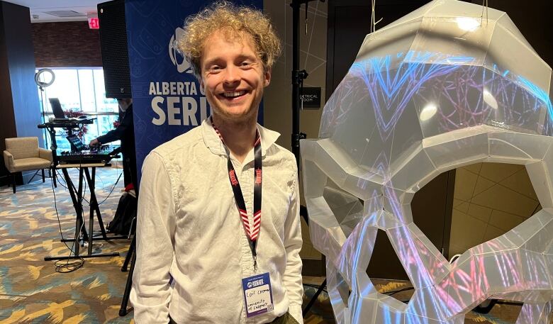 26-year-old Loc Cremer stands in front of a blue-purple vertical banner advertising the Alberta Games Series. His curly blonde hair sits messily as he smiles wide for the photo. 