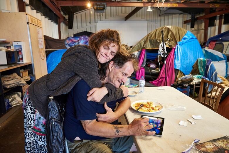 A woman leans over to hug a friend who is sitting at a table playing with an iPad.