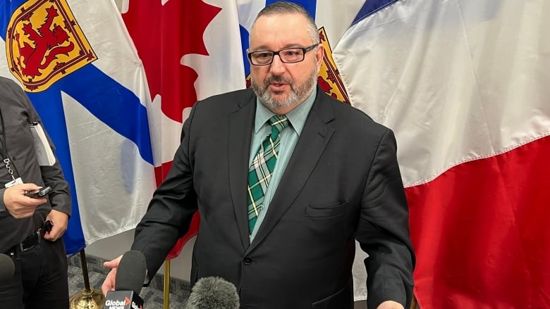 A man with glasses and a beard talks to reporters with outstretched microphones.