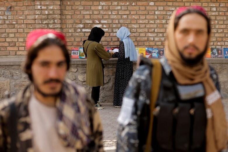 Two men are seen in the foreground. Two girls, wearing head coverings, are seen in the background facing a brick wall and looking at books on a table.