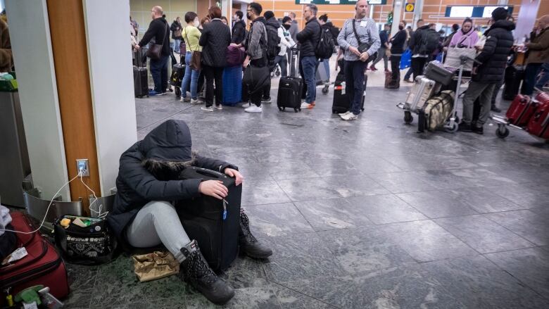 A passenger with their hood pulled up sits on the floor and leans against a pole with their head bowed at Vancouver International Airport.