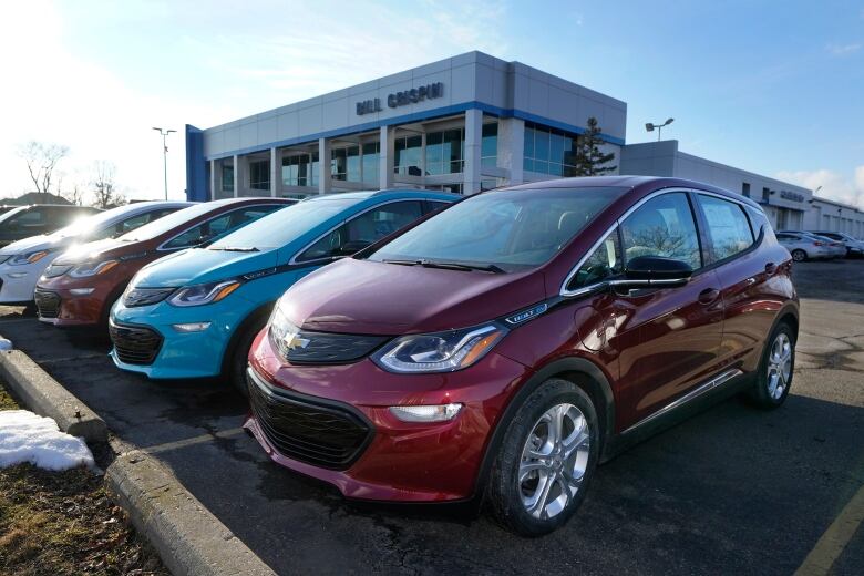 Red and blue cars in the parking lot outside a white dealership building.