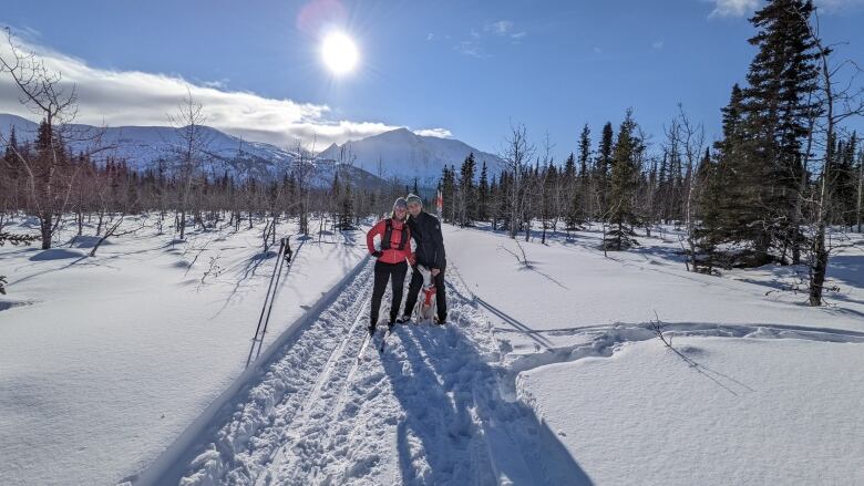 A woman and I man stand on a snowy trail with a dog between them. It's sunny and there's snow-covered mountains in the background. They are both dressed in winter coats and hats, and are smiling.