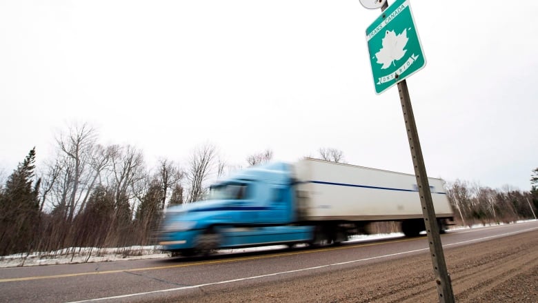 A semi-truck drives along a highway in front of a road sign reading 