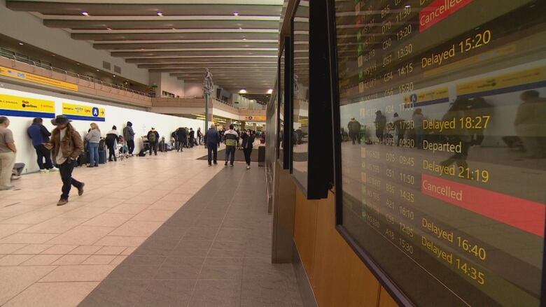 People line up against a wall. A screen is shown on the side of the image, with several flights showing they're delayed or cancelled.