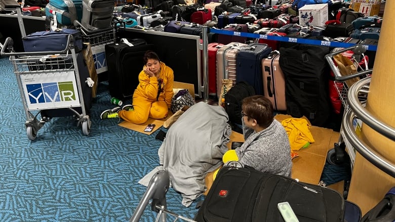 Two Filipina women sit on cardboard on the floor of an airport, with roped-off luggage behind them. Someone is lying between them, covered in a grey blanket.