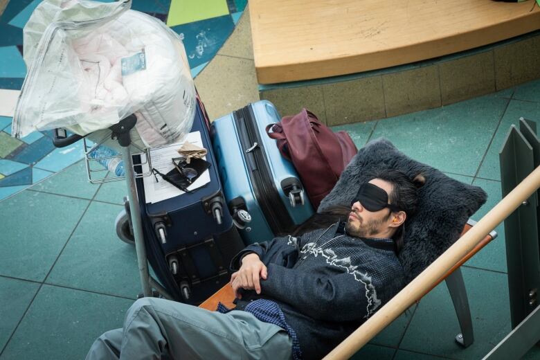 A bearded man sleeps next to a pile of luggage with a black eye mask covering his eyes and bright orange earplugs in his ears.