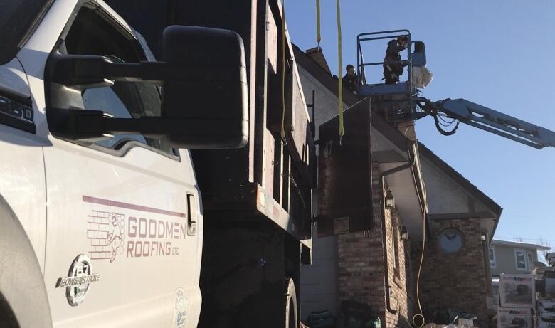 A vehicle with a Goodmen Roofing logo is parked next to a house where employees are replacing a roof.