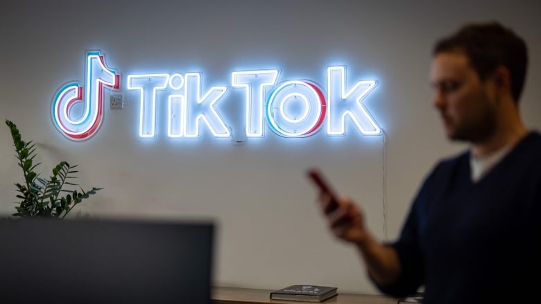 A man, who is out of focus, looks at his cellphone while standing near a neon 'TikTok' sign.