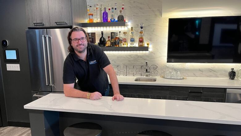 A man leans over a counter in his basement suite. In the background, there's a fridge, a tv and about a dozen alcohol bottles lined up on shelves.