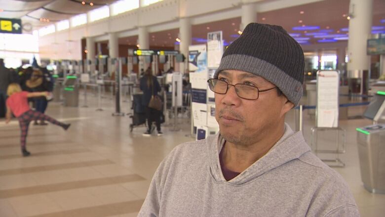 A man with glasses and wearing a dark grey toque is standing in the airport departures terminal.