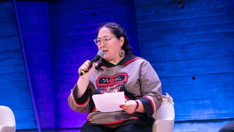 Aluki Kotierk sit on stage, speaking into a microphone. She wears beaded earrings and a matching beaded shirt. 