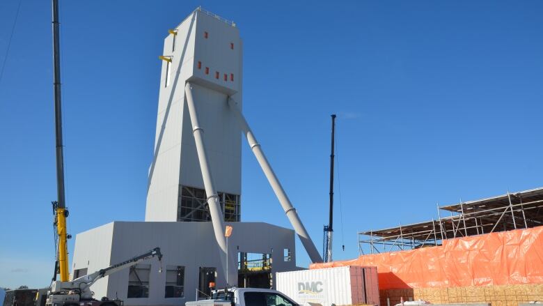 Cranes and other construction equipment surround a large grey structure with two big legs and square windows at the top arranged in the shape of a smiley face. 