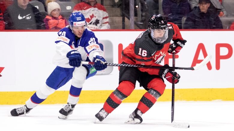 A Canadian hockey player is to the right side of the picture wearing red and black gear. He keeps the puck from an Slovakian player, wearing blue and white gear, as some members of the audience watch from behind the glass.