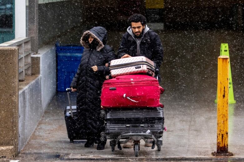 A couple with several pieces of luggage are seen walking outside an airport terminal as it's snowing.