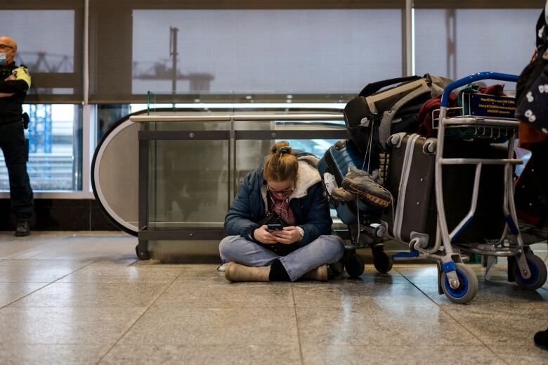 A woman sits cross-legged on the floor, looking at her cellphone next to a cart full of luggage.