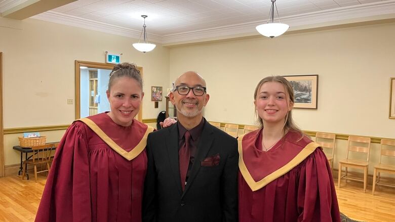 Two women dressed in red robes and gold trim, one man dressed in a black suit with glasses and a maroon tie, are standing inside a spacious community room.