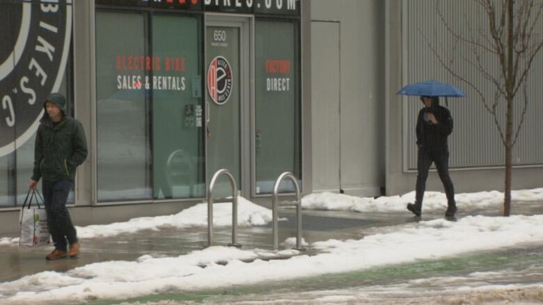 Two people cross a pavement with piles of snow around them. One of them is holding an umbrella, and both are wearing multiple layers.