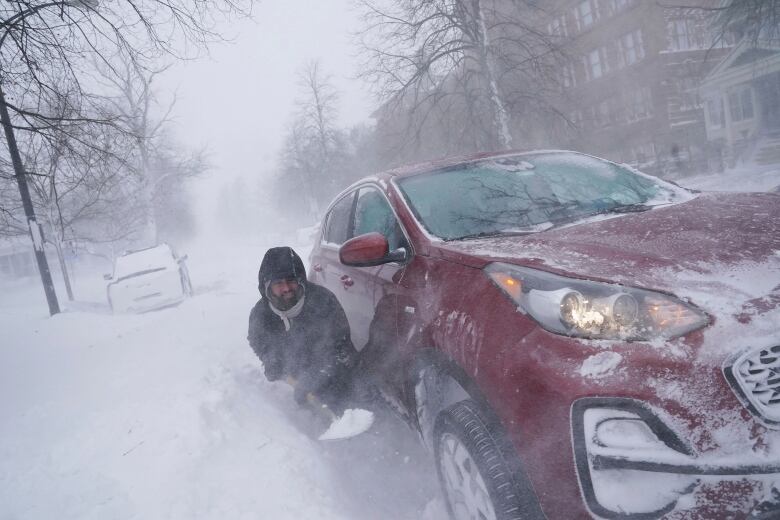 A man shovels snow beside a red car in a snowstorm.