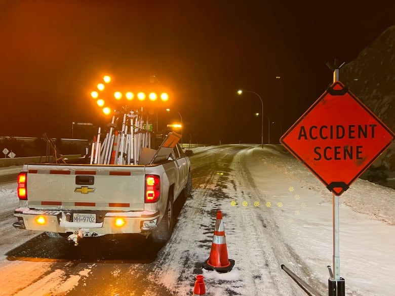 A pickup truck with a flashing leftward arrow parked on a snowy highway. A sign next to it reads 'ACCIDENT SCENE'. Orange traffic cones are visible.