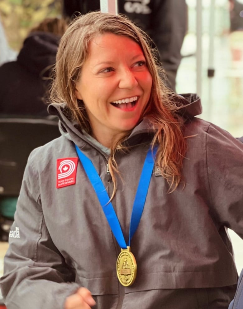 A woman with wet hair wearing a jacket smiles while wearing a gold medal with a blue ribbon around her neck.