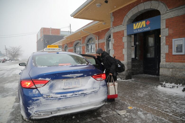 A person in a coat and mask speaks with the driver of a dark blue taxi outside a Via Rail station on a snowy day.