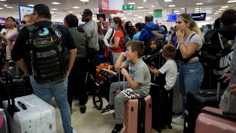 A large number of people, many wearing backpacks, stand around luggage. Some children are sitting on suitcases.