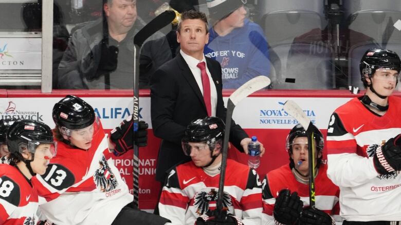 A head coach in a suit looks skyward behind hockey players on the bench in white jerseys, one of whom has one leg over the boards.