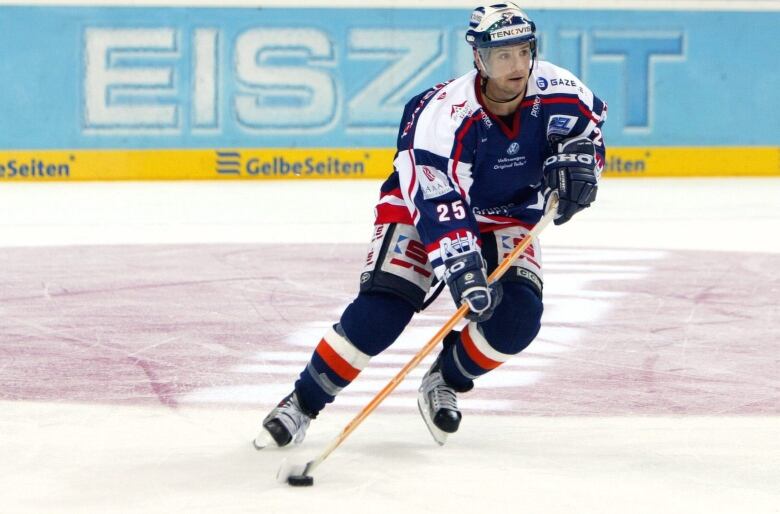 A hockey player dressed in a blue, red and white uniform skates with the puck on the ice.