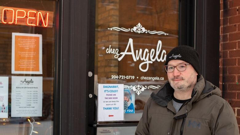 A man wearing a big winter jacket and toque stands outside a door that says 