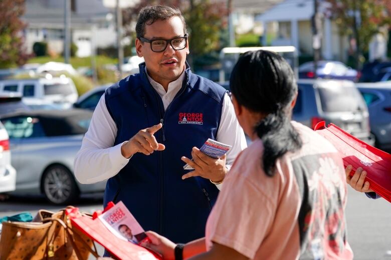 A man in a blue vest, white shirt and glasses speaks with a person whose back is to the camera.