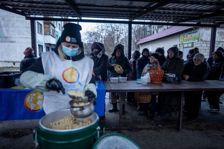 A person spoons food from a large pot into a smaller container as people line up behind a table.