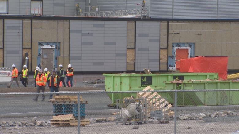 A group of construction workers in bright vests and hard hats walk in front of a new building that is under construction.