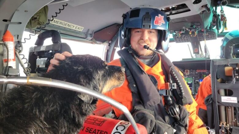 A man wearing high-vis clothing and seated in a helicopter with equipment pets a dog that sits on a seat nearby.