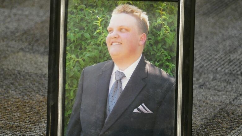 A framed photo of a blonde man smiling in a suit and tie sits on a carpeted floor.