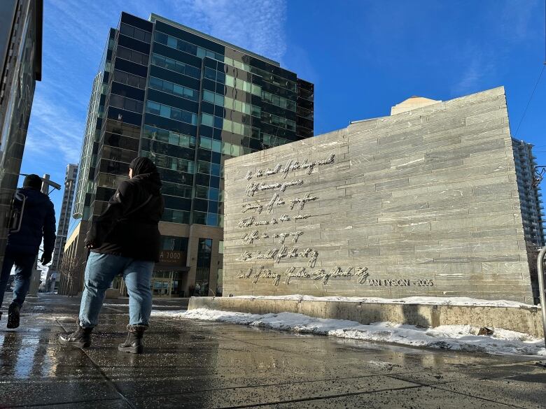 A woman walks before a grey wall with metal engravings of lyrics in Ian Tyson's handwriting. 